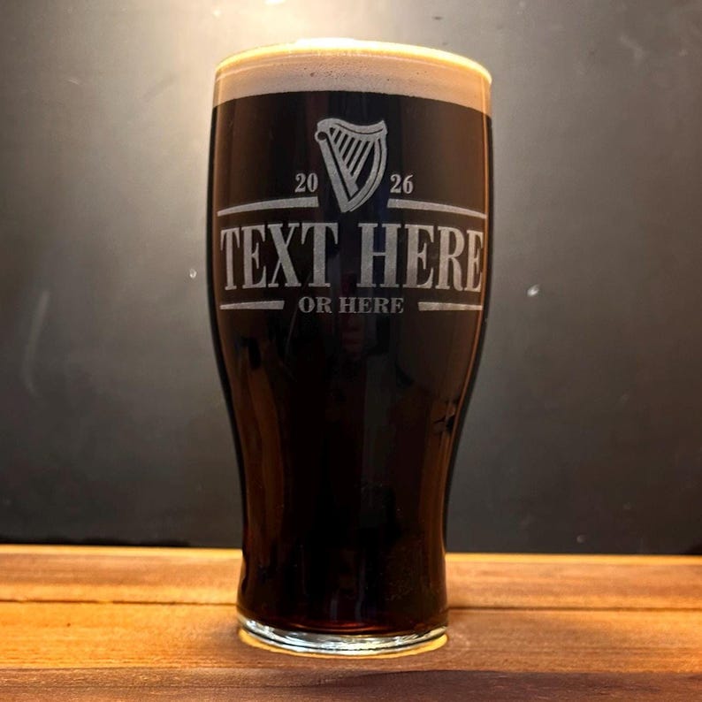 May include: A pint glass filled with dark beer and a creamy head. The glass has etched text that reads "TEXT HERE" and "OR HERE" below a harp symbol and the numbers "20" and "26". The glass is on a wooden surface.