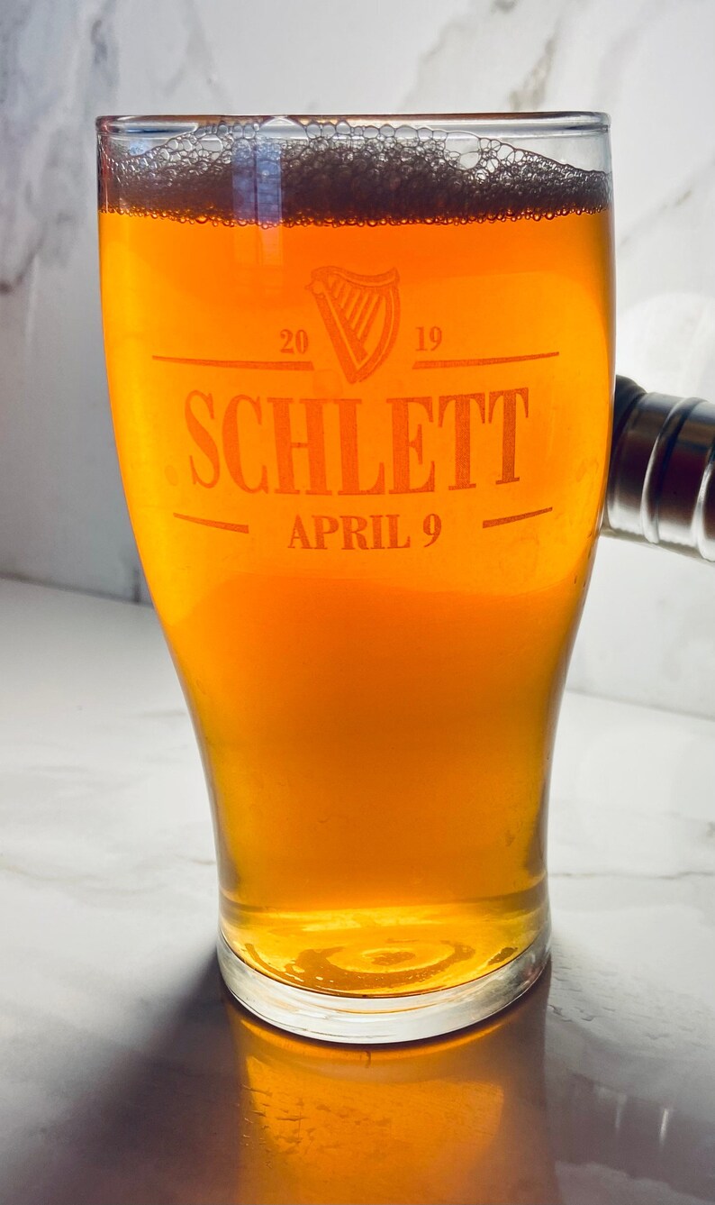 May include: A glass of amber-colored beer with a white head in a clear glass pint glass. The glass has the text "Schlett" and "April 9" etched into it.