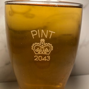 May include: A clear glass pint glass with the words "PINT" and "2043" etched on the side. A crown with a design inside is etched between the words.