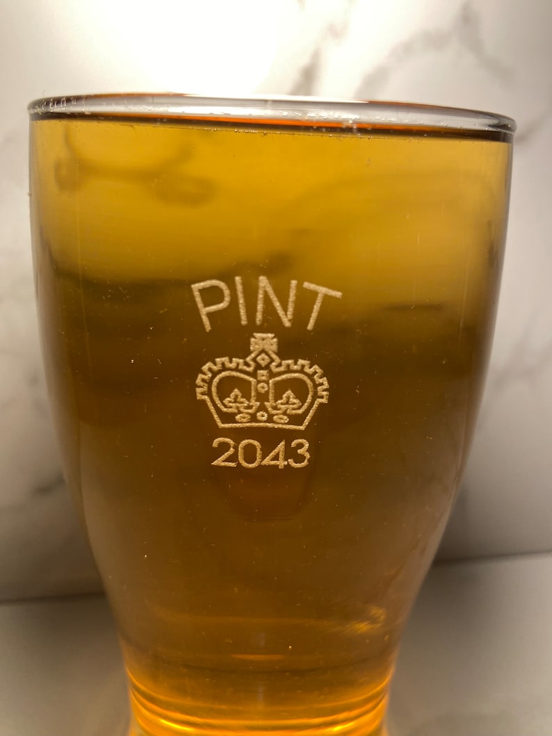 May include: A clear glass pint glass with the words "PINT" and "2043" etched on the side. A crown with a design inside is etched between the words.