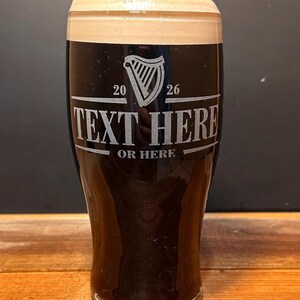 May include: A pint glass filled with dark beer and a creamy head. The glass has etched text that reads "TEXT HERE" and "OR HERE" with a harp symbol and the numbers "20" and "26". The glass is on a wooden surface.