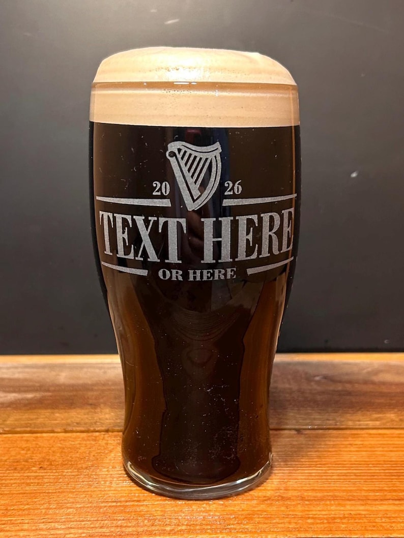 May include: A pint glass filled with dark beer and a creamy head. The glass has etched text that reads "TEXT HERE" and "OR HERE" with a harp symbol and the numbers "20" and "26". The glass is on a wooden surface.