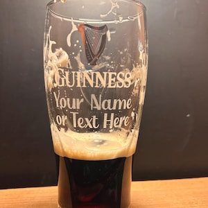 May include: A pint glass with the Guinness logo and the text "Your Name or Text Here". The glass is partially filled with a dark stout, topped with a creamy head. The glass has a classic pint shape, ideal for enjoying a pint.