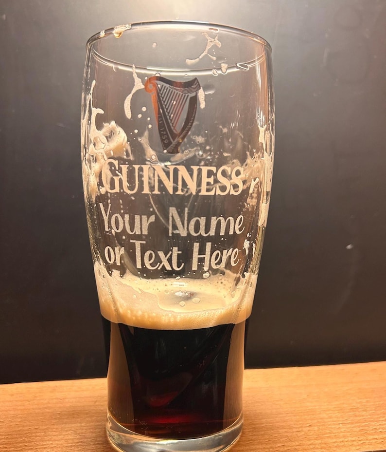 May include: A pint glass with the Guinness logo and the text "Your Name or Text Here". The glass is partially filled with a dark stout, topped with a creamy head. The glass has a classic pint shape, ideal for enjoying a pint.