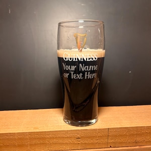 May include: A pint glass filled with dark stout beer and a creamy head. The glass is etched with the word "GUINNESS" and the text "Your Name or Text Here". The glass is on a wooden surface.