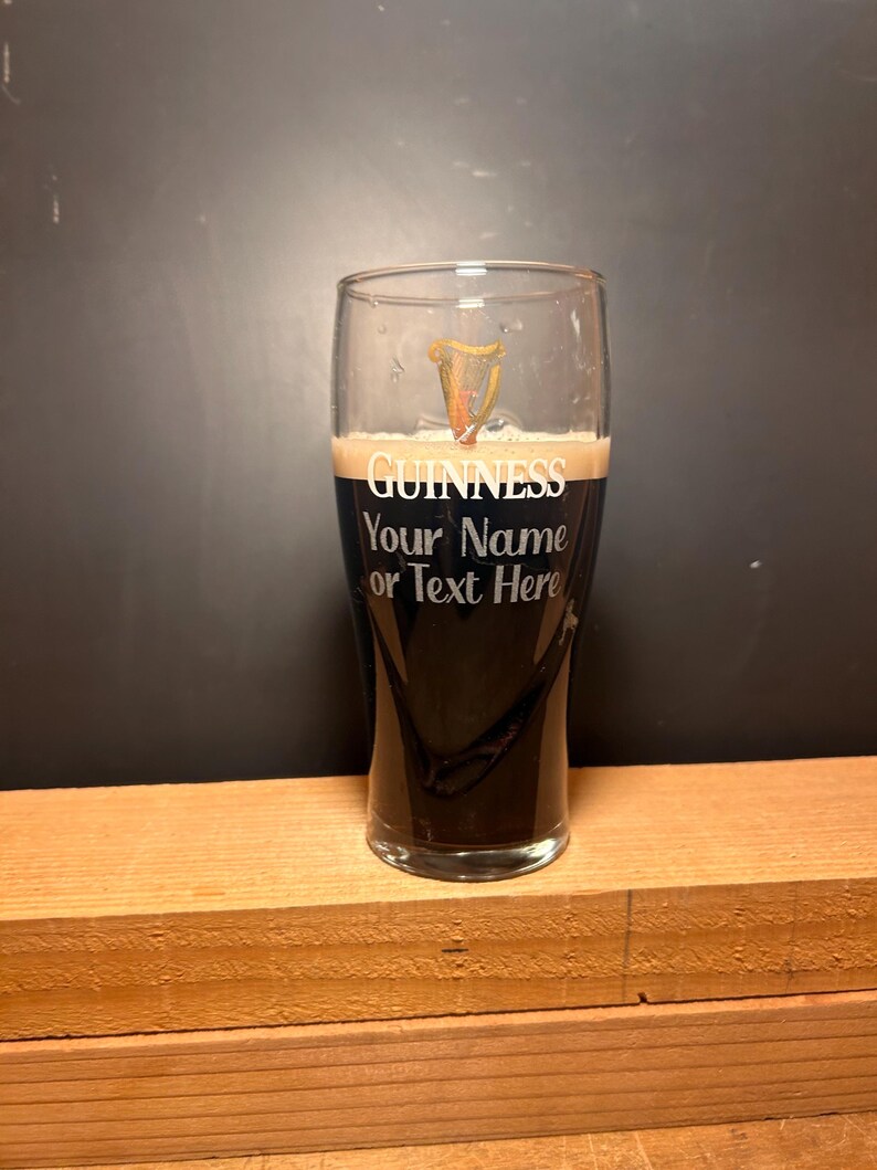 May include: A pint glass filled with dark stout beer and a creamy head. The glass is etched with the word "GUINNESS" and the text "Your Name or Text Here". The glass is on a wooden surface.