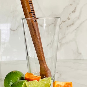 May include: A wooden cocktail muddler with the text "Gramppy's" engraved on it, sitting in a clear glass.  There are lime and orange slices on a white marble surface.