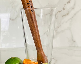 May include: A wooden cocktail muddler with the text "Gramppy's" engraved on it, sitting in a clear glass.  There are lime and orange slices on a white marble surface.