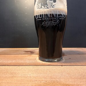 May include: A pint glass of dark stout beer with a thick, creamy white head. The glass is branded with the Guinness logo and the word "Draught". The glass is on a wooden surface.