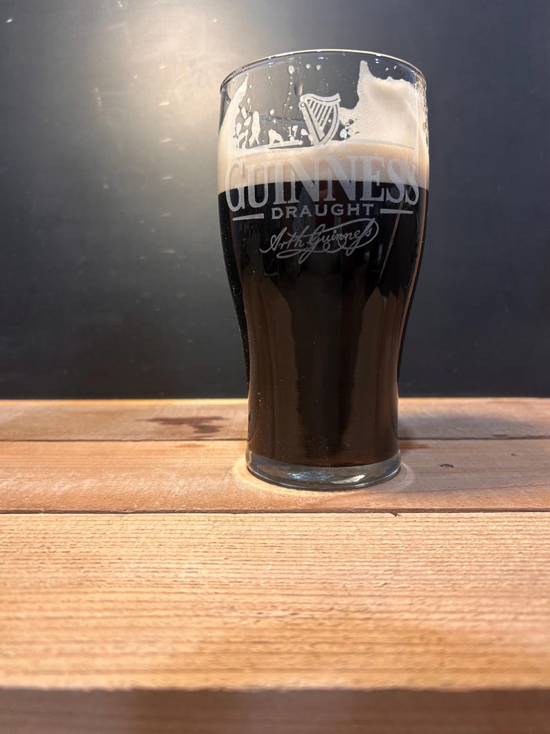 May include: A pint glass of dark stout beer with a thick, creamy white head. The glass is branded with the Guinness logo and the word "Draught". The glass is on a wooden surface.