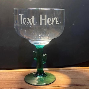 May include: A clear glass goblet with a green cactus-shaped stem and base. The words "Text Here" are etched in white on the glass. The goblet sits on a wooden surface.