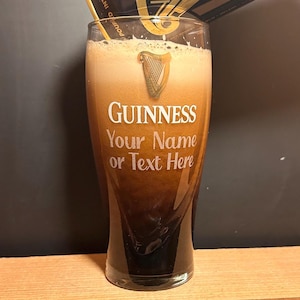 May include: A pint glass filled with dark stout and a creamy head. The glass has the Guinness logo and the text "Your Name or Text Here". The glass is on a wooden surface. The beer is a rich, dark brown colour.