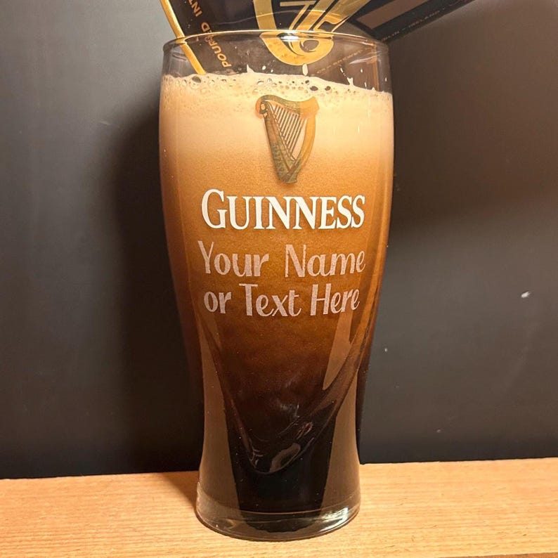 May include: A pint glass filled with dark stout and a creamy head. The glass has the Guinness logo and the text "Your Name or Text Here". The glass is on a wooden surface. The beer is a rich, dark brown colour.
