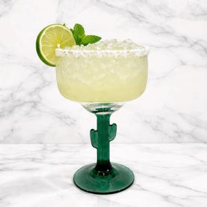 Pu&ograve; includere: Un cocktail margarita in un bicchiere verde a forma di cactus, guarnito con una fetta di lime e un rametto di menta. Il bicchiere ha un bordo salato e poggia su una superficie di marmo bianco. La bevanda &egrave; di colore giallo chiaro.