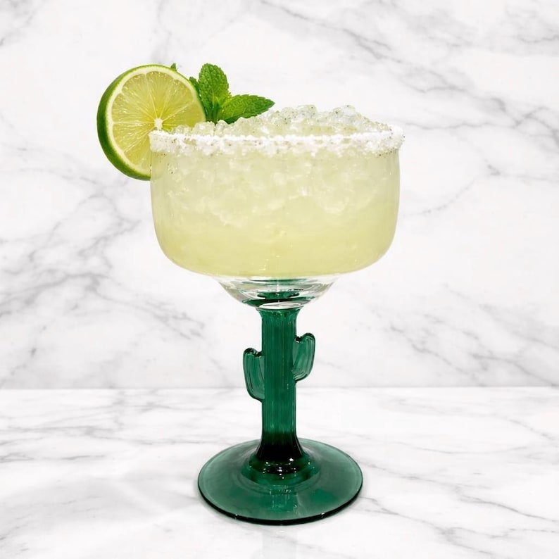 Pu&ograve; includere: Un cocktail margarita in un bicchiere verde a forma di cactus, guarnito con una fetta di lime e un rametto di menta. Il bicchiere ha un bordo salato e poggia su una superficie di marmo bianco. La bevanda &egrave; di colore giallo chiaro.
