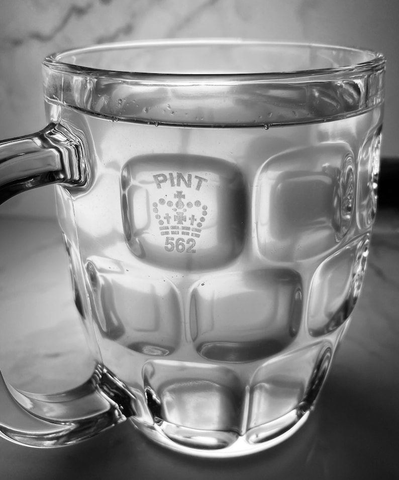 K&ouml;nnte beinhalten: Ein klares Glas-Bierglas mit einer strukturierten Oberfl&auml;che. Das Glas hat eine Krone und den Text "PINT 562" auf der Seite.