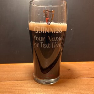 May include: A pint glass filled with dark stout, displaying the Guinness logo and the text "Your Name or Text Here." The glass has a harp emblem. The glass is on a wooden surface.