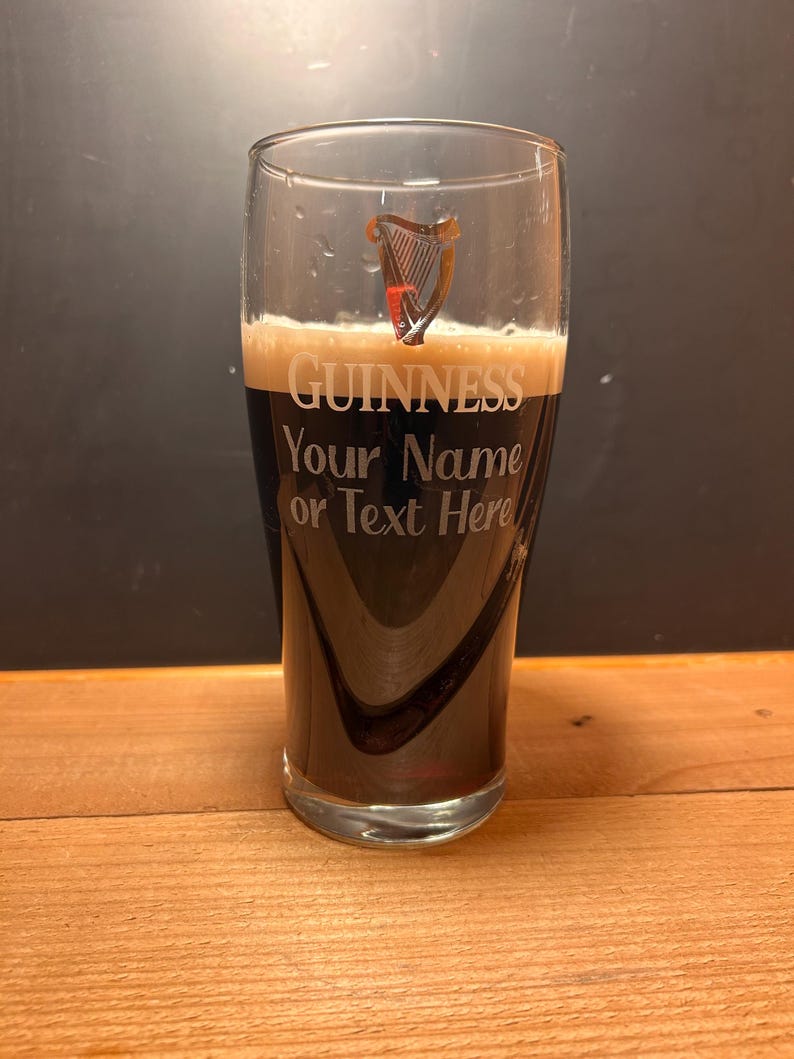 May include: A pint glass filled with dark stout, displaying the Guinness logo and the text "Your Name or Text Here." The glass has a harp emblem. The glass is on a wooden surface.