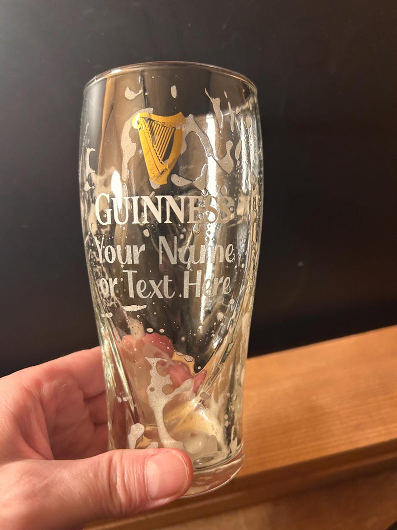 May include: A pint glass with the Guinness logo and the text "Your Name or Text Here". The glass has beer residue and foam. The Guinness logo is a gold harp.