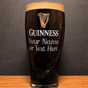 May include: A tall, dark Guinness pint glass with a creamy head. The glass features the Guinness logo, a golden harp, and the text "GUINNESS Your Name or Text Here" in white. The glass is on a wooden surface.