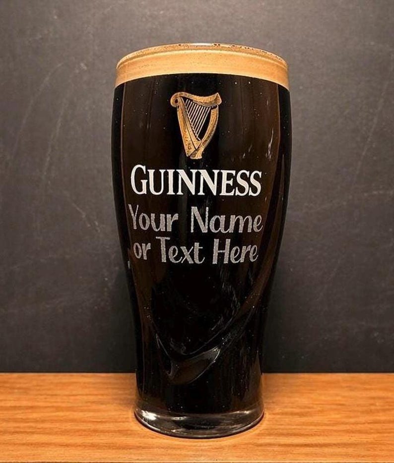 May include: A black Guinness pint glass with a tan head. The glass has the Guinness logo, a golden harp, and the text "GUINNESS Your Name or Text Here" in white. The glass is on a wooden surface.