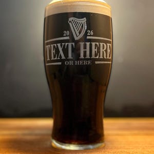 May include: A pint glass filled with dark stout beer, topped with a creamy white head. The glass has etched text that reads "TEXT HERE" and "OR HERE" below a harp symbol and the numbers "20" and "26". The glass sits on a wooden surface.