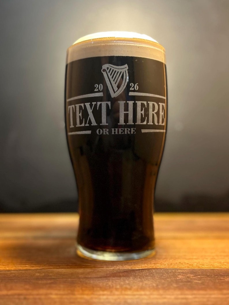 May include: A pint glass filled with dark stout beer, topped with a creamy white head. The glass has etched text that reads "TEXT HERE" and "OR HERE" below a harp symbol and the numbers "20" and "26". The glass sits on a wooden surface.