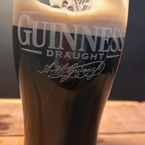 May include: A pint glass of Guinness Draught, a dark stout. The glass is etched with the Guinness logo, including the words "Guinness Draught" and "Arth Guinness". The beer has a creamy, tan head, contrasting with the dark liquid.