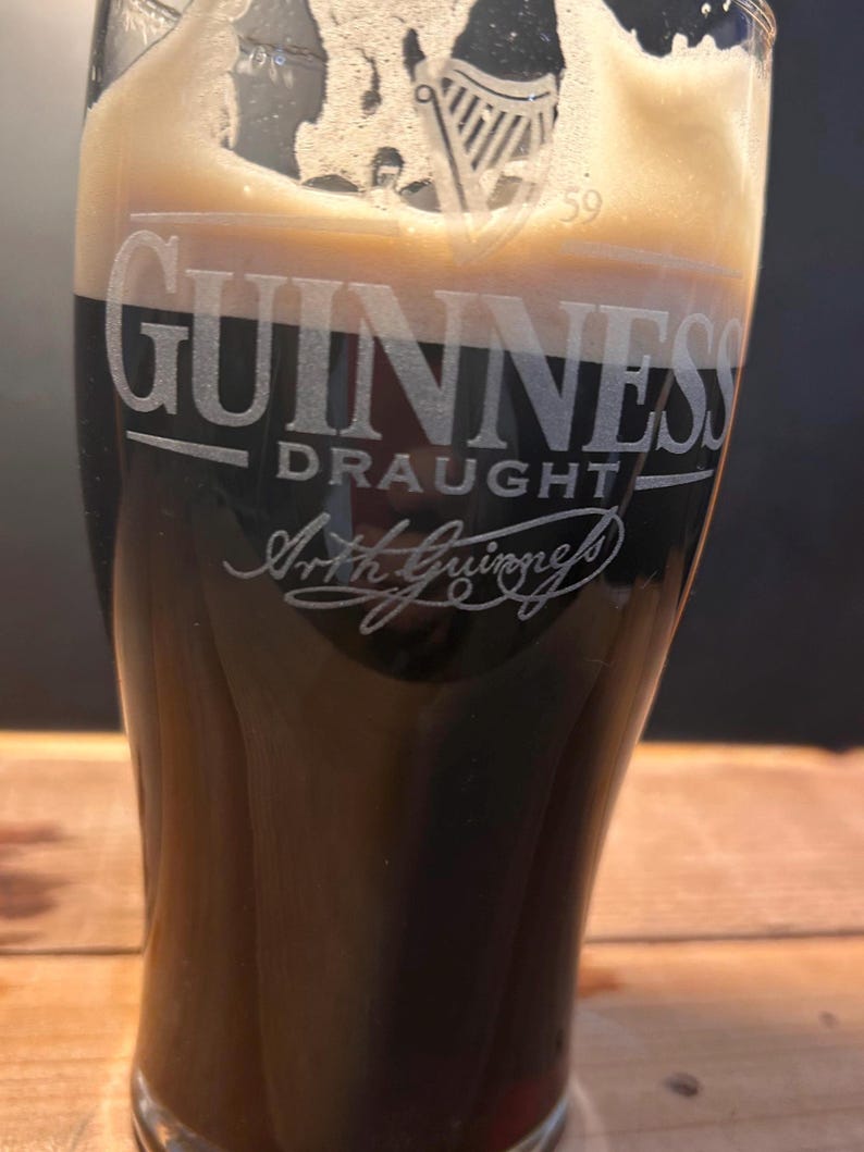May include: A pint glass of Guinness Draught, a dark stout. The glass is etched with the Guinness logo, including the words "Guinness Draught" and "Arth Guinness". The beer has a creamy, tan head, contrasting with the dark liquid.