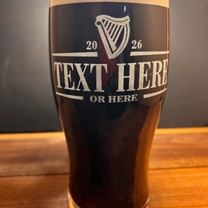 May include: A pint glass filled with dark beer and a creamy head. The glass has a harp logo and the text "TEXT HERE OR HERE" etched on the front. The glass sits on a wooden surface.