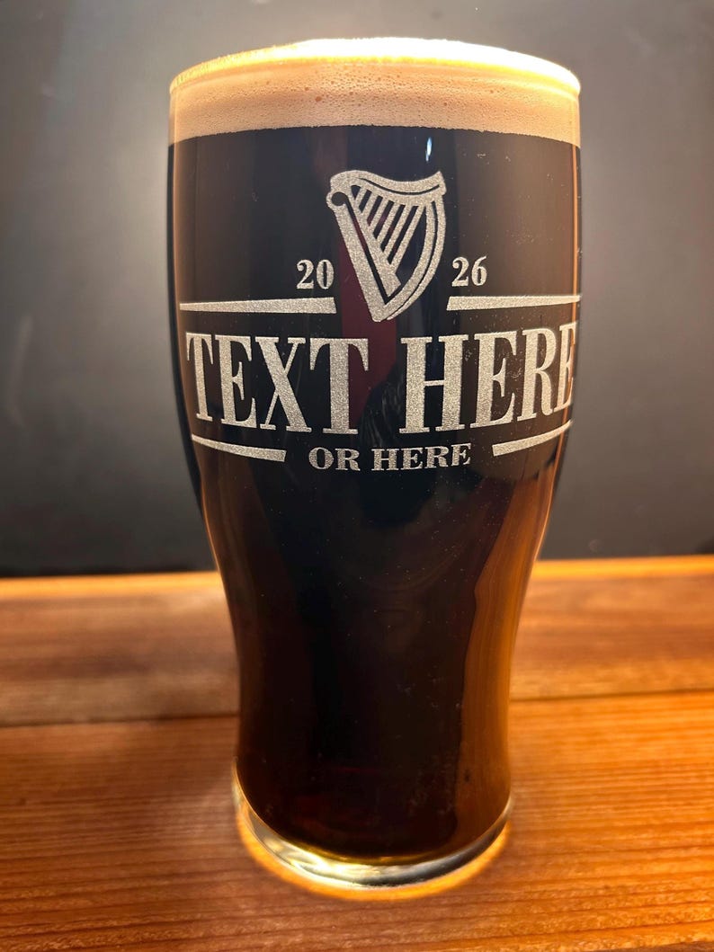 May include: A pint glass filled with dark beer and a creamy head. The glass has a harp logo and the text "TEXT HERE OR HERE" etched on the front. The glass sits on a wooden surface.