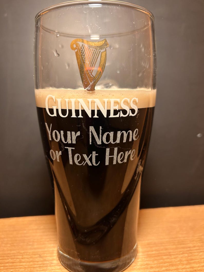 May include: A pint glass filled with dark stout beer, topped with a creamy head. The glass features the Guinness logo and the text "Your Name or Text Here". The Guinness logo is a gold harp.