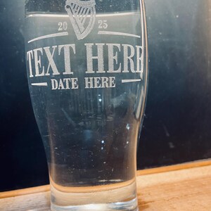 May include: A clear glass pint glass with an etched design. The design includes a harp, the text "TEXT HERE", and the text "DATE HERE".