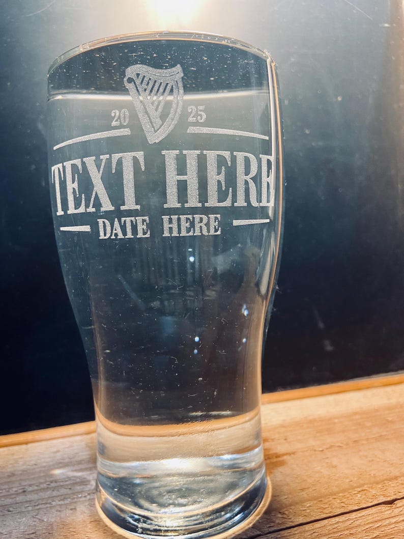 May include: A clear glass pint glass with an etched design. The design includes a harp, the text "TEXT HERE", and the text "DATE HERE".