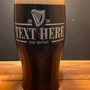 May include: A pint glass filled with dark beer and a creamy head. The glass is etched with a harp symbol, the numbers 20 and 26, and the text "TEXT HERE OR HERE" in a rectangular frame. The glass sits on a wooden surface.