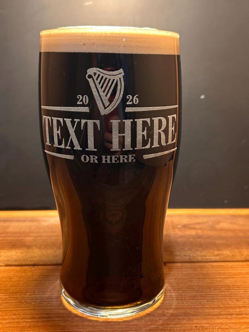 May include: A pint glass filled with dark beer and a creamy head. The glass is etched with a harp symbol, the numbers 20 and 26, and the text "TEXT HERE OR HERE" in a rectangular frame. The glass sits on a wooden surface.