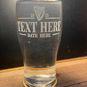May include: A clear glass pint glass with an etched design. The design includes a harp, the numbers 20 and 25, the text "TEXT HERE", and the text "DATE HERE".