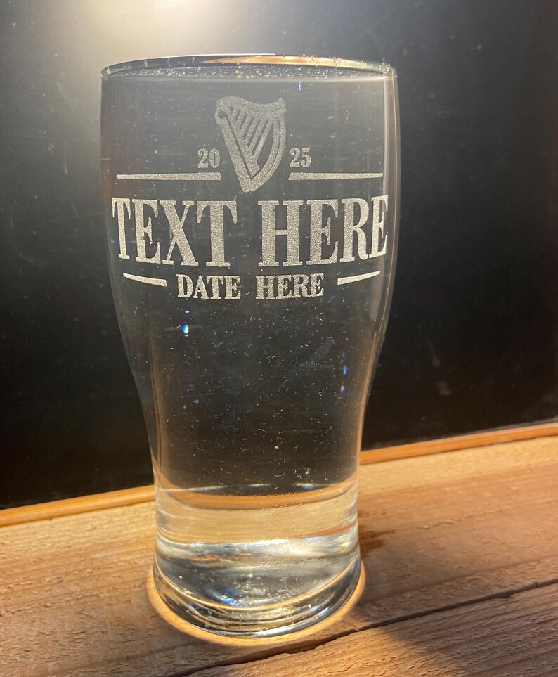 May include: A clear glass pint glass with an etched design. The design includes a harp, the numbers 20 and 25, the text "TEXT HERE", and the text "DATE HERE".