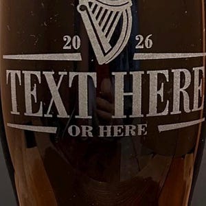 May include: A dark pint glass with a white harp logo and the text "TEXT HERE OR HERE". The numbers "20" and "26" are on either side of the harp. The glass is filled with a dark liquid and topped with a foamy head.