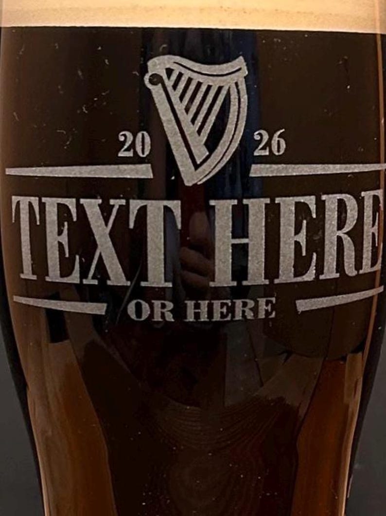 May include: A dark pint glass with a white harp logo and the text "TEXT HERE OR HERE". The numbers "20" and "26" are on either side of the harp. The glass is filled with a dark liquid and topped with a foamy head.