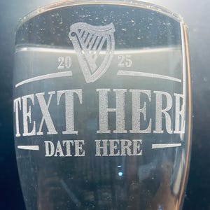 May include: A clear glass pint glass with an etched design. The design includes a harp, the numbers 20 and 25, the text "TEXT HERE", and the text "DATE HERE".