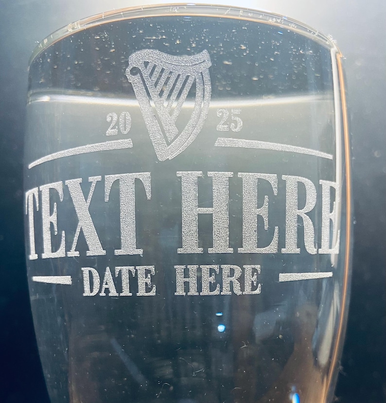 May include: A clear glass pint glass with an etched design. The design includes a harp, the numbers 20 and 25, the text "TEXT HERE", and the text "DATE HERE".