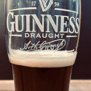 May include: A pint glass of dark stout with a thick, creamy white head. The glass is branded with the Guinness logo, a white harp, and the words "GUINNESS DRAUGHT" and "Arth Guinness" in white.