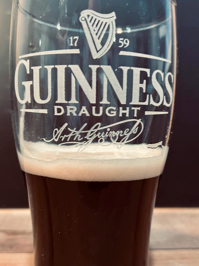 May include: A pint glass of dark stout with a thick, creamy white head. The glass is branded with the Guinness logo, a white harp, and the words "GUINNESS DRAUGHT" and "Arth Guinness" in white.