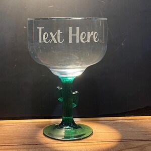 May include: A clear margarita glass with a green cactus stem and base. The text "Text Here" is etched on the glass. The glass sits on a wooden surface against a dark background.