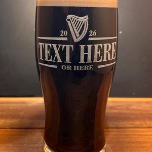 May include: A tall, dark glass of stout beer with a thick, creamy head. The glass is etched with a harp design and the text "TEXT HERE OR HERE". The glass is on a wooden surface.