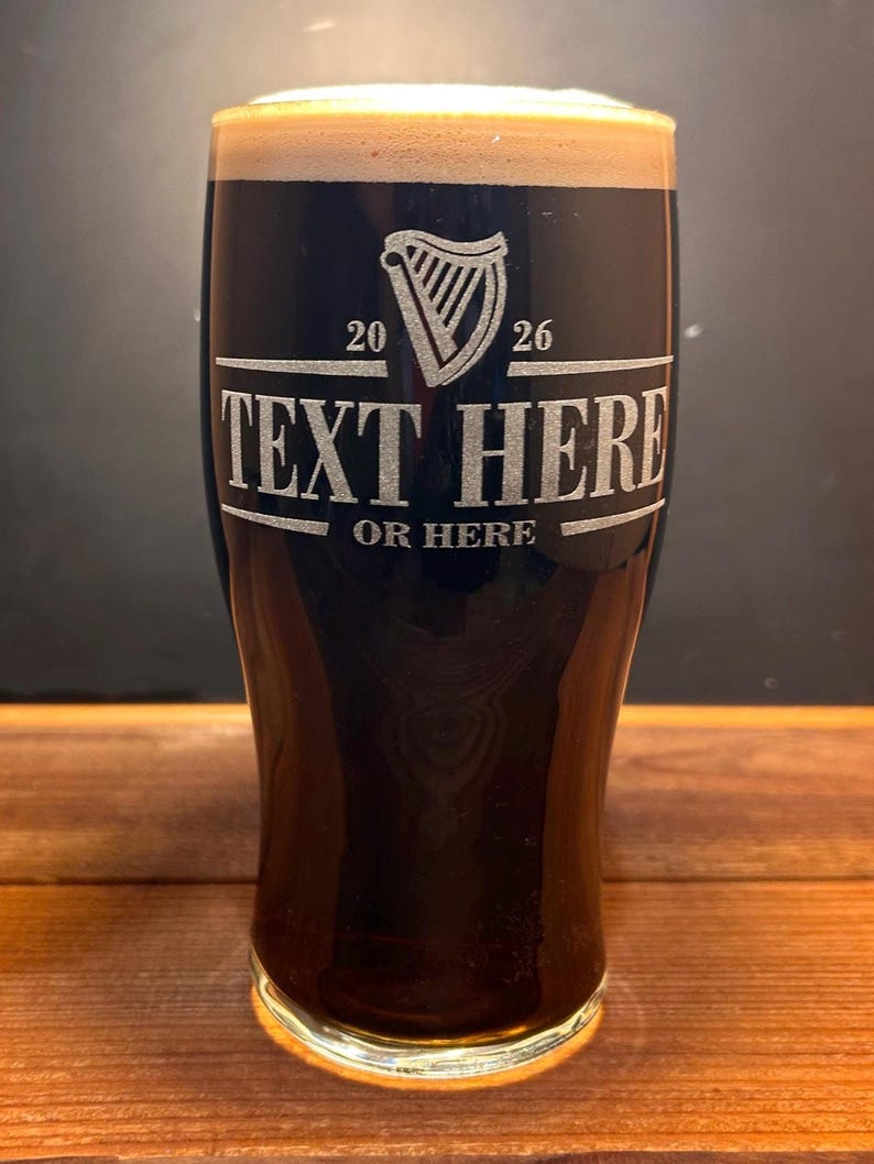 May include: A tall, dark glass of stout beer with a thick, creamy head. The glass is etched with a harp design and the text "TEXT HERE OR HERE". The glass is on a wooden surface.