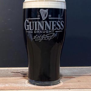 May include: A pint glass of Guinness stout with a thick, creamy head. The glass features the Guinness logo, including the harp symbol and the words "Guinness Draught". The glass is set on a wooden surface, with a dark background.