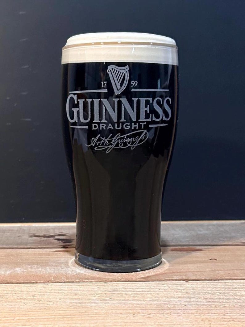 May include: A pint glass of Guinness stout with a thick, creamy head. The glass features the Guinness logo, including the harp symbol and the words "Guinness Draught". The glass is set on a wooden surface, with a dark background.