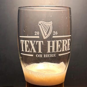May include: A clear pint glass with a white harp logo, the numbers 20 and 26, and the words "TEXT HERE" and "OR HERE" etched on the front. The glass is partially filled with a dark beer and foam.